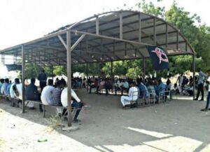 Ungefähr 50 Menschen versammeln sich an der Universität von Dongola unter einer Überdachung. Einige hören zu von der Seite. In der Mitte hängt eine anarchistische Fahne.